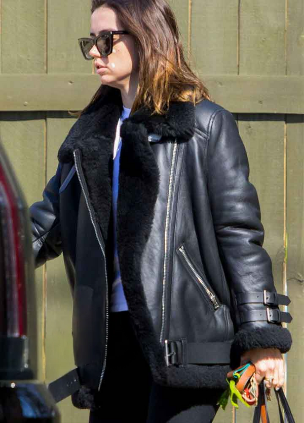 Person wearing a black leather jacket with fur trim, standing against a green wooden wall.